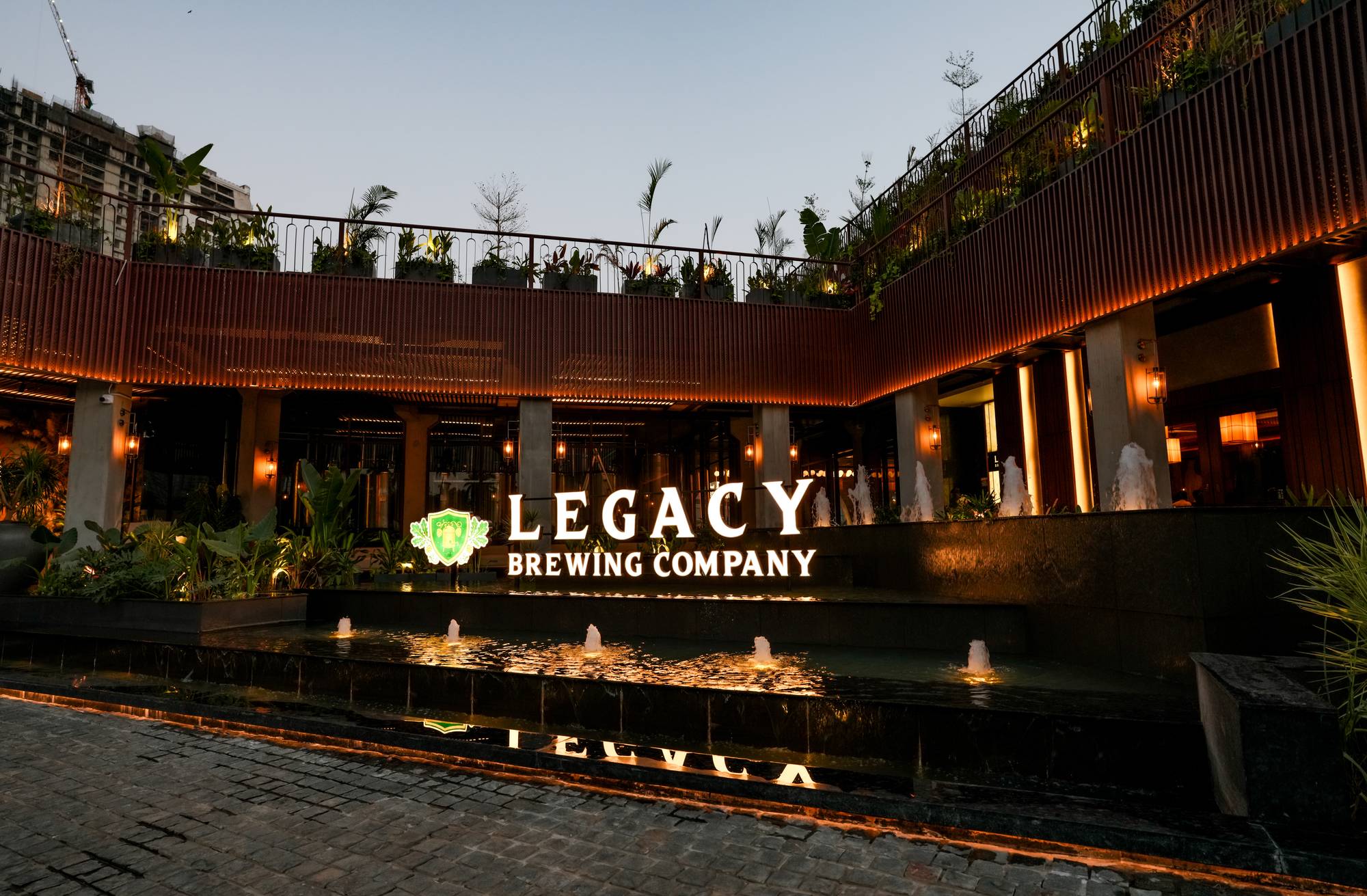 Legacy Brewing Company exterior signage lit up at dusk beside the fountain plaza